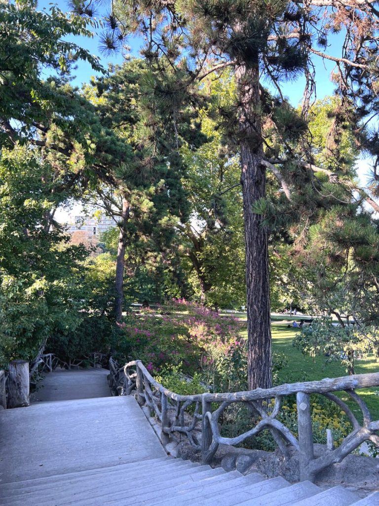 Eine breite Steintreppe mit rustikalen Holzgeländern führt durch einen grünen Park voller hoher Bäume und blühender Sträucher. Das Sonnenlicht scheint durch das Blätterdach. Parc Montouris