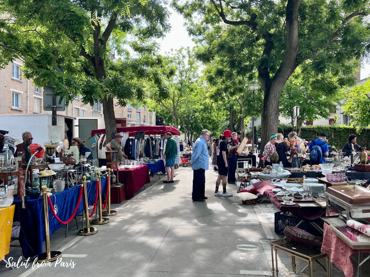 Marché aux Puces de Vanves mit Verkaufstischen links und rechts und grünen Bäumen. 