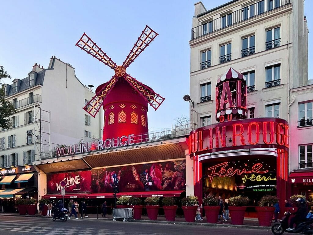 Moulin Rouge mit roter Windmühle und leuchtenden Neonlichtern bei Dämmerung mit Menschen davor. Dieses berühmte Kabarett ist eine lebendige Paris Sehenswürdigkeiten Attraktion.