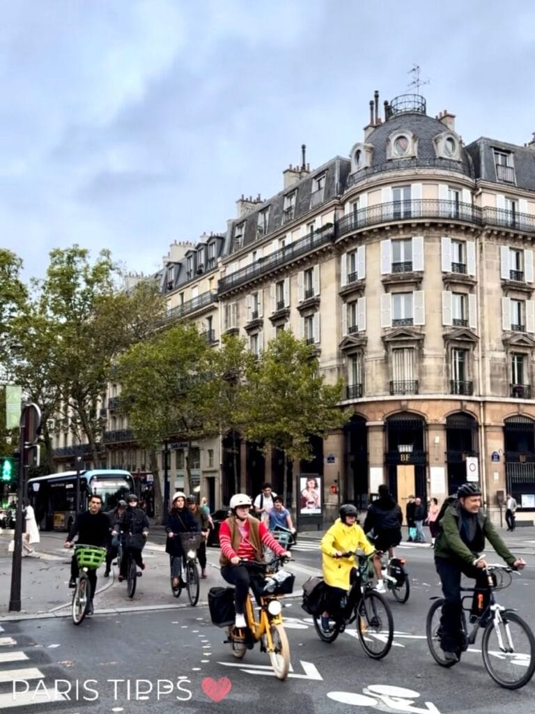 Viele Radfahrer überqueren eine Kreuzung in Paris auf einem stark genutzten Radweg. Die Szene zeigt, wie intensiv der Radverkehr inzwischen ist und dass die Fahrradwege oft wie Verkehrsachsen genutzt werden. Typisch für das moderne Radfahren in Paris.
