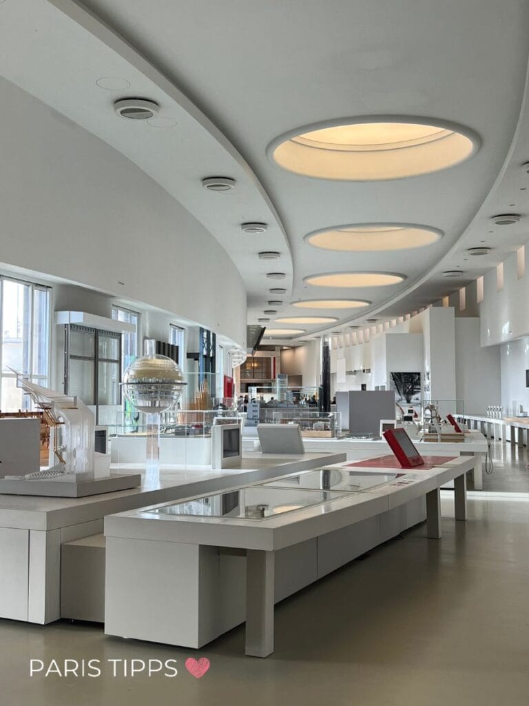Innenansicht der Cité de l’Architecture et du Patrimoine in Paris. Der moderne Ausstellungsraum zeigt Architekturmodelle, Pläne und interaktive Stationen auf weißen Tischen. Die Decke ist geschwungen mit großen, runden Lichtfeldern, die warmes Licht spenden. Im Hintergrund sind weitere Modelle und Glasvitrinen zu sehen. Die Atmosphäre ist hell, ruhig und futuristisch.