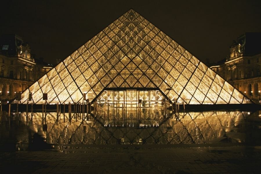 Die gläserne Pyramide des Louvre-Museums bei Nacht, beeindruckend beleuchtet und gespiegelt im umgebenden Wasser. Die goldene Beleuchtung hebt die Struktur hervor, während die historischen Gebäude des Louvre im Hintergrund im Dunkeln liegen.