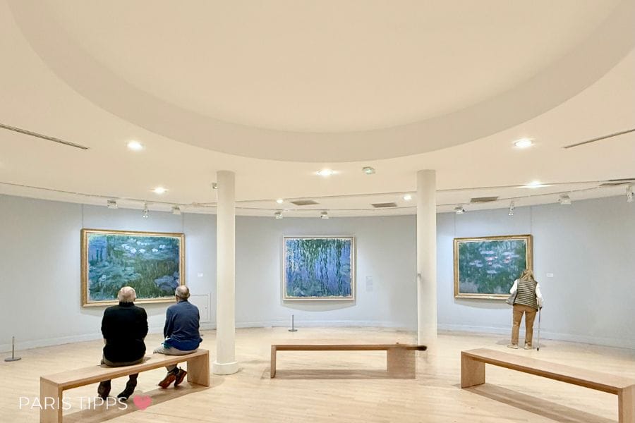 Ein heller Ausstellungsraum im Musée Marmottan Monet in Paris. An den Wänden hängen mehrere großformatige Gemälde von Claude Monet, darunter seine berühmten Seerosen und Gartenmotive. Zwei Personen sitzen auf einer Holzbank und betrachten die Kunstwerke in Ruhe, eine weitere Person steht nah vor einem Bild. Der Raum ist schlicht und rund angelegt, mit hellem Holzboden und weichem Licht.