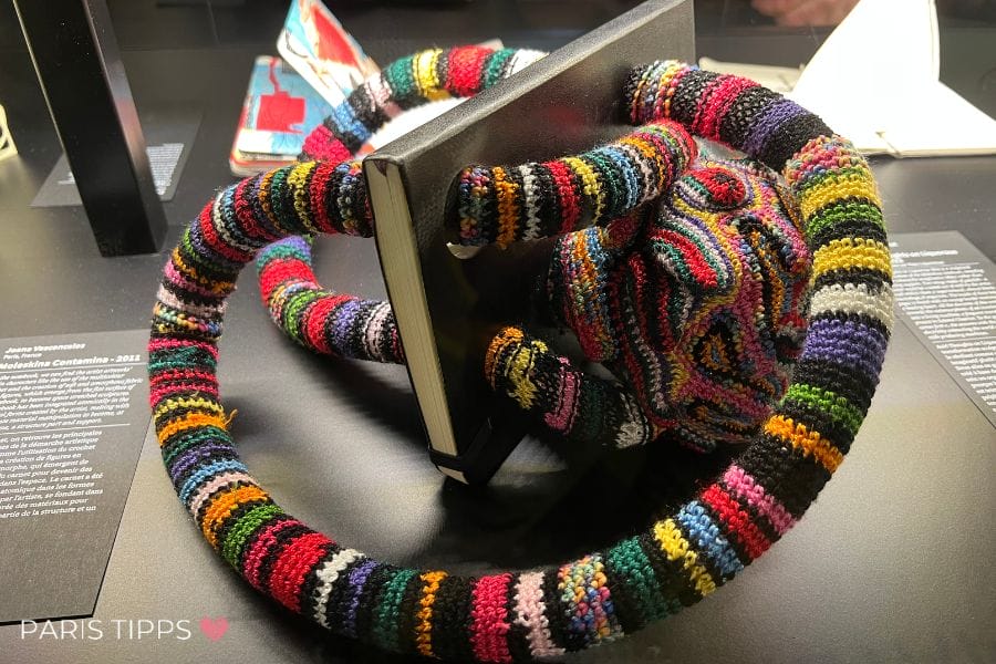 Bunte, gehäkelte Skulptur in Tentakel-Form, die ein schwarzes Notizbuch umschlingt – modernes Kunstobjekt aus dem Palais de Tokyo in Paris.