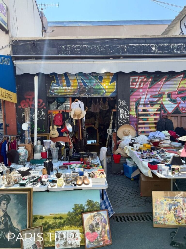 Flohmarktstand auf den Puces de Montreuil in Paris mit Tischen voller Vintage-Objekte. Zu sehen sind Schmuck, Geschirr, Bilderrahmen, alte Lampen und verschiedene Gebrauchsgegenstände. Vor dem Stand hängen Kleidung und Hüte. Im Hintergrund sieht man Graffiti an der Hauswand. Die Szene zeigt das typische, etwas chaotische Flair dieses Pariser Flohmarkts.