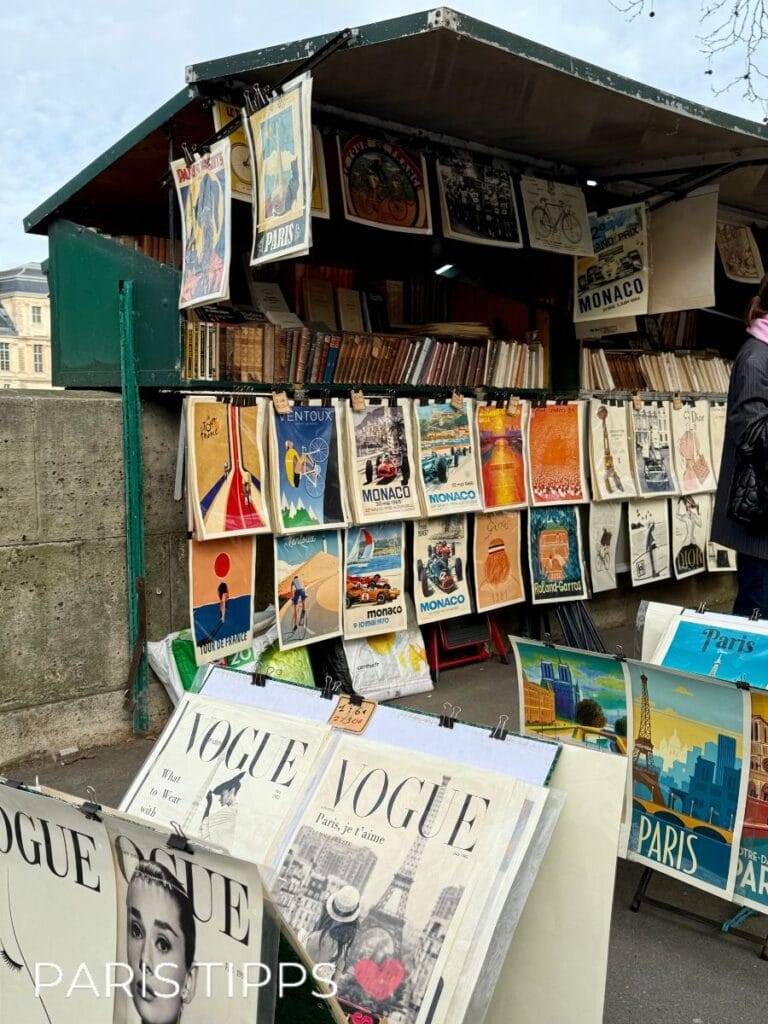 Bouquinistes-Stand an der Seine in Paris mit typischen grünen Bücherkisten. Ausgestellt sind Vintage-Poster, alte Magazine und Bücher, darunter mehrere Ausgaben der Modezeitschrift Vogue. Zwischen den Drucken hängen klassische Reiseplakate von Paris und Monaco. Der Stand zeigt das charakteristische Bild der Pariser Buchhändler entlang der Seineufer.