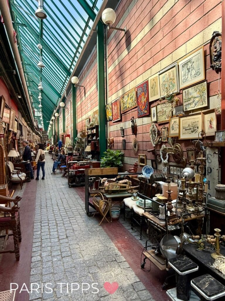 Überdachte Passage des Flohmarkts Saint-Ouen in Paris mit Antiquitätenständen entlang eines gepflasterten Ganges. Händler präsentieren Möbel, Lampen, Bilderrahmen und Vintage-Dekoration vor einer roten Backsteinwand. Besucher schlendern zwischen den Ständen. Das Glasdach darüber lässt viel Licht hinein und zeigt die typische Atmosphäre der Pariser Flohmarkthallen.