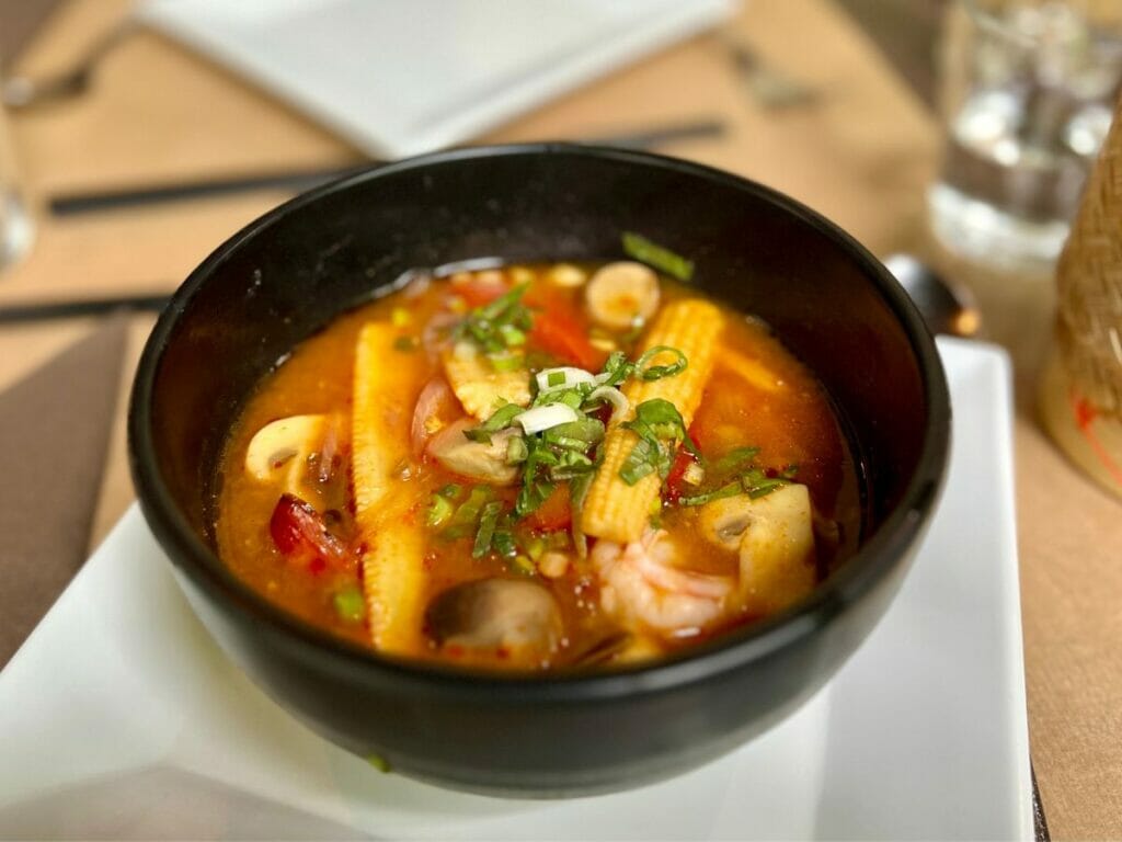Lao Lane Xang, hier Eine dampfende Schüssel mit asiatischer Suppe, gefüllt mit Babymais, Pilzen, Garnelen, Frühlingszwiebeln und roter Paprika. Die Suppe steht auf einem weißen Teller in einem Restaurant. 