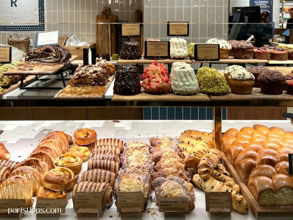 Paris Geheimtipps: Leckeres Gebäck in Lafayette Gourmet Eine bunte Auswahl an französischem Gebäck, darunter Schokoladenkuchen, Pistazienmuffins und Croissants, präsentiert in einer eleganten Bäckerei – ein süßer Paris Geheimtipp für Feinschmecker.