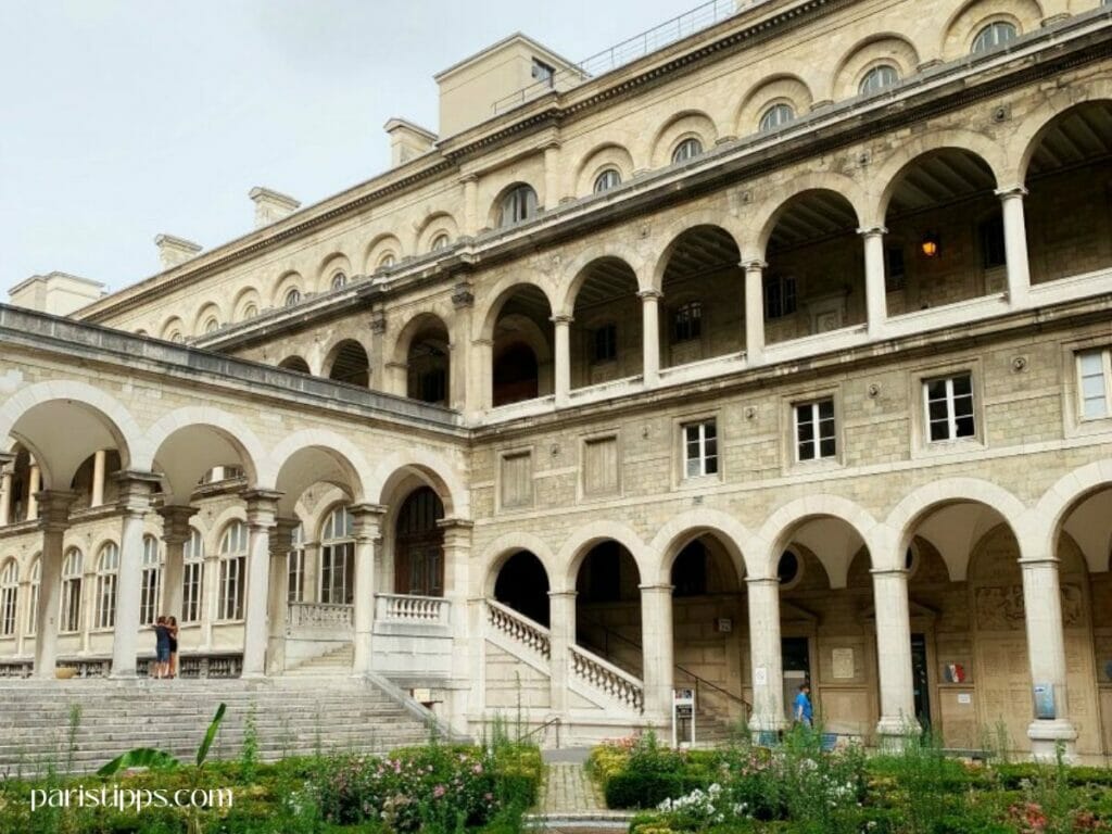 Historischer Gebäudekomplex mit Rundbögen, Säulen und Treppenaufgang, umgeben von einem gepflegten Garten – ein architektonischer Paris Geheimtipp in der Nähe von Notre-Dame.