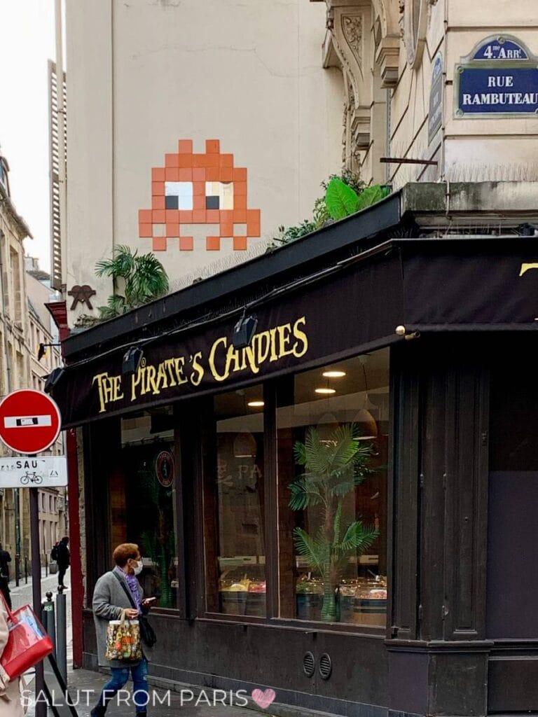Space Invader in Orange an Hausmauer in Paris. Darunter ein Süßigkeitenladen und ein Einbahnstrassenschild. 