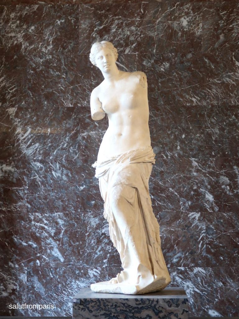 Die berühmte Statue der Venus von Milo im Louvre-Museum in Paris. Die antike Marmorfigur steht auf einem marmorgemusterten Sockel vor einer dunkel marmorierten Wand. Die Skulptur zeigt eine weibliche Figur mit unbekleidetem Oberkörper und fehlenden Armen. Der Raum ist ruhig und elegant inszeniert.
