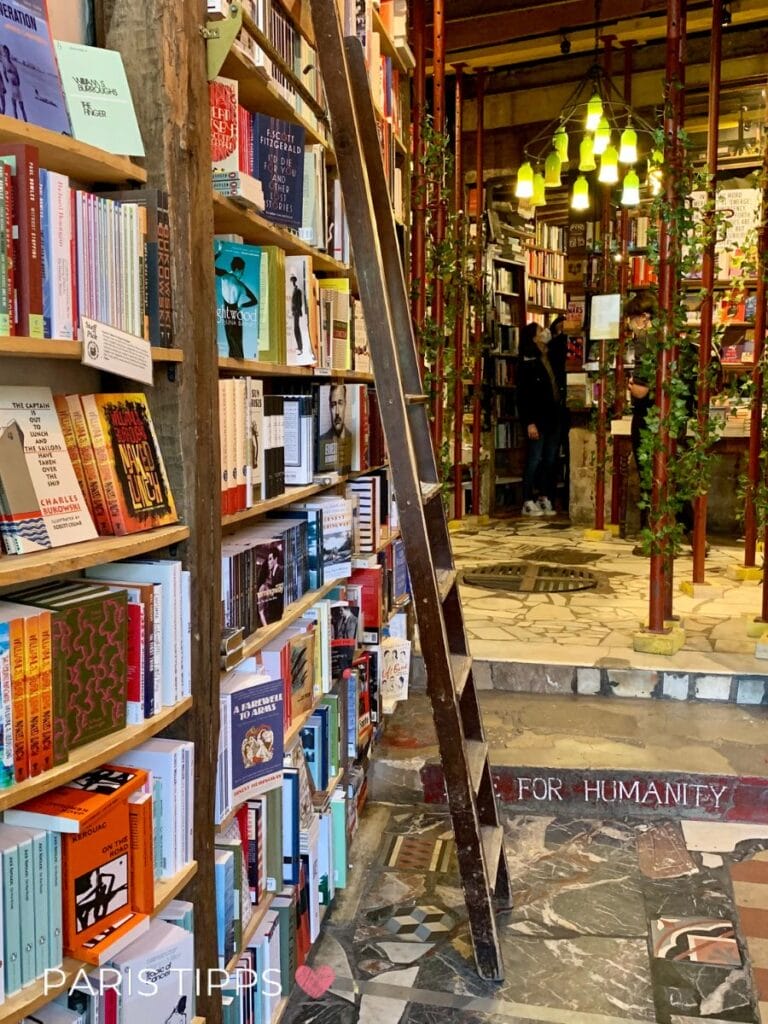 Innenraum eines Buchladens mit Regalen voller Bücher und einer Leiter sowie dem Text "FOR HUMANITY" auf einer Stufe. "PARIS TIPPS ♥" steht unten im Bild und zeigt einen gemütlichen Ort in Paris im Regen.
