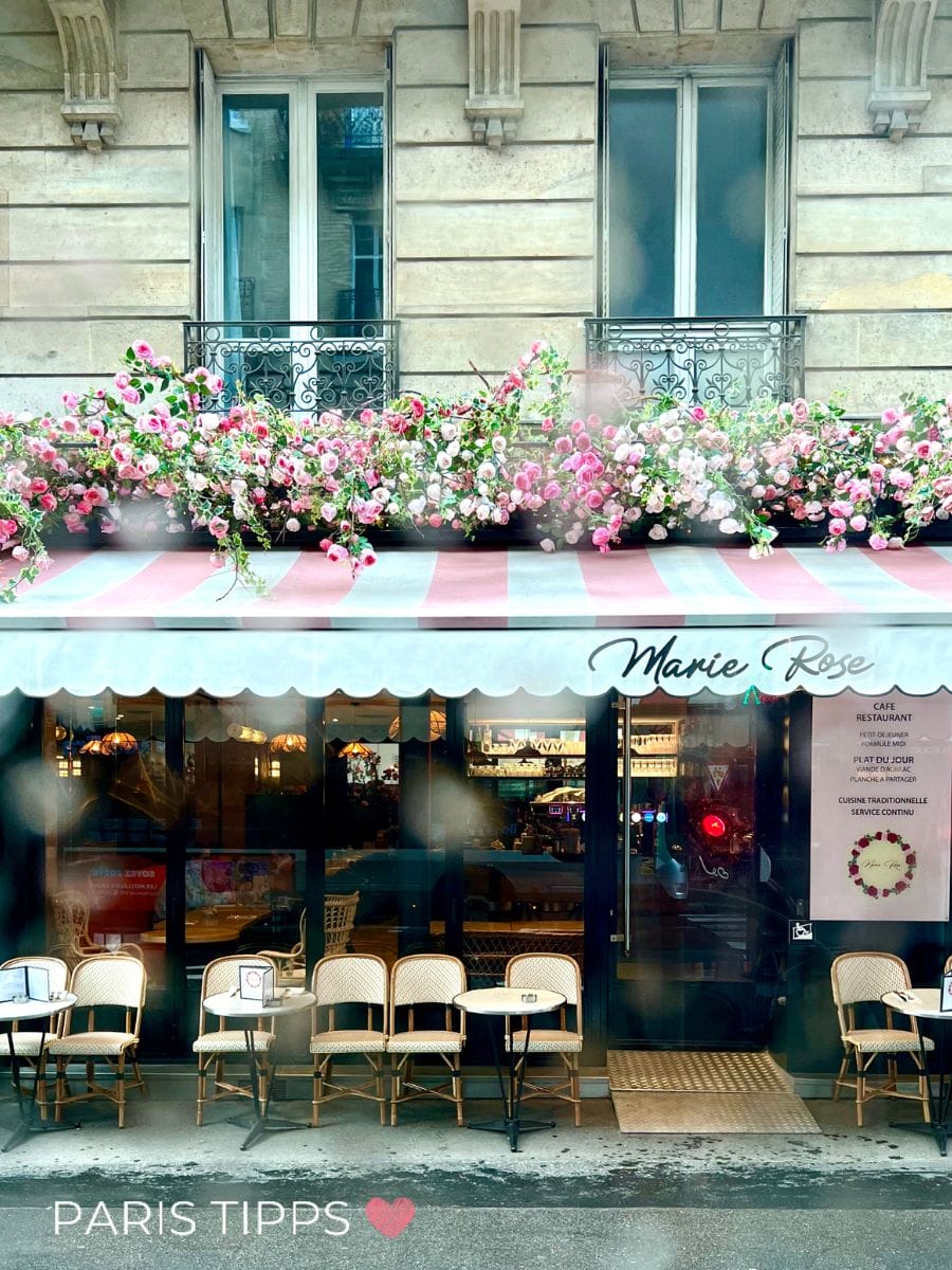 Café mit Blumendekoration über der Markise und Sitzplätzen draußen in Paris im Regen. Das Schild zeigt "Marie Rose" sowie Menütexte wie "CAFE RESTAURANT" und "CUISINE TRADITIONNELLE SERVICE CONTINU".