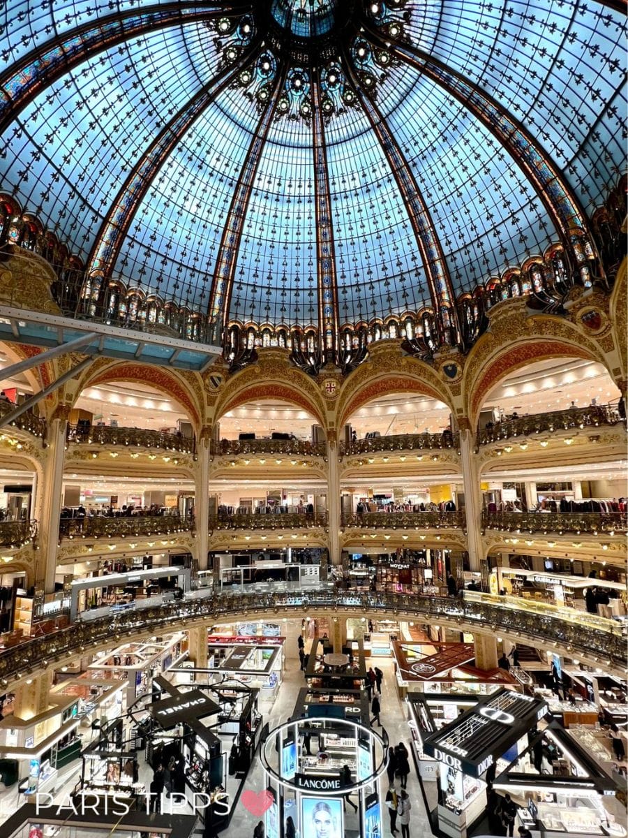 Innenansicht eines großen Kaufhauses mit gläserner Kuppel und mehreren Etagen voller Geschäfte in Paris im Regen. "PARIS TIPPS ♥" steht unten im Bild und zeigt Shopping in Paris im Regen.