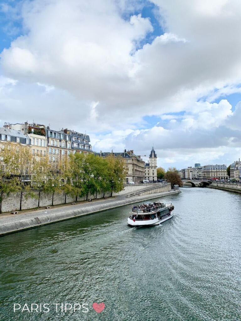 Boot fährt auf der Seine vorbei an historischen Gebäuden unter bewölktem Himmel in Paris im Regen. "PARIS TIPPS ♥" steht unten im Bild und zeigt eine Aussicht auf Paris an einem grauen vom Wasser aus.