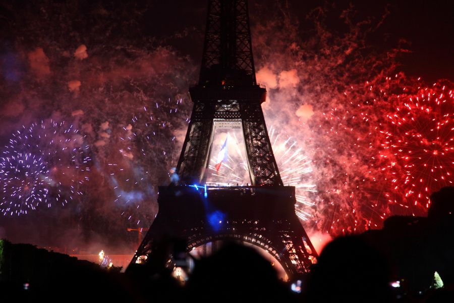 Ein großes Feuerwerk erhellt den Nachthimmel rund um den Eiffelturm in Paris, mit roten, weißen und blauen Farben. Die französische Flagge wird auf den Turm projiziert. Zahlreiche Zuschauer haben sich versammelt, um das Ereignis zu verfolgen – eine Kombination aus Lichtershow, nationaler Symbolik und urbaner Kulisse.
