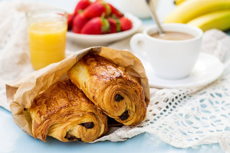 2 Pain au Chocolat in einer braunen Papiertüte und im Hingrund verschwommen einen Tasse Milchcafe, orangensaft und ein schälchen ERdbeeren.
