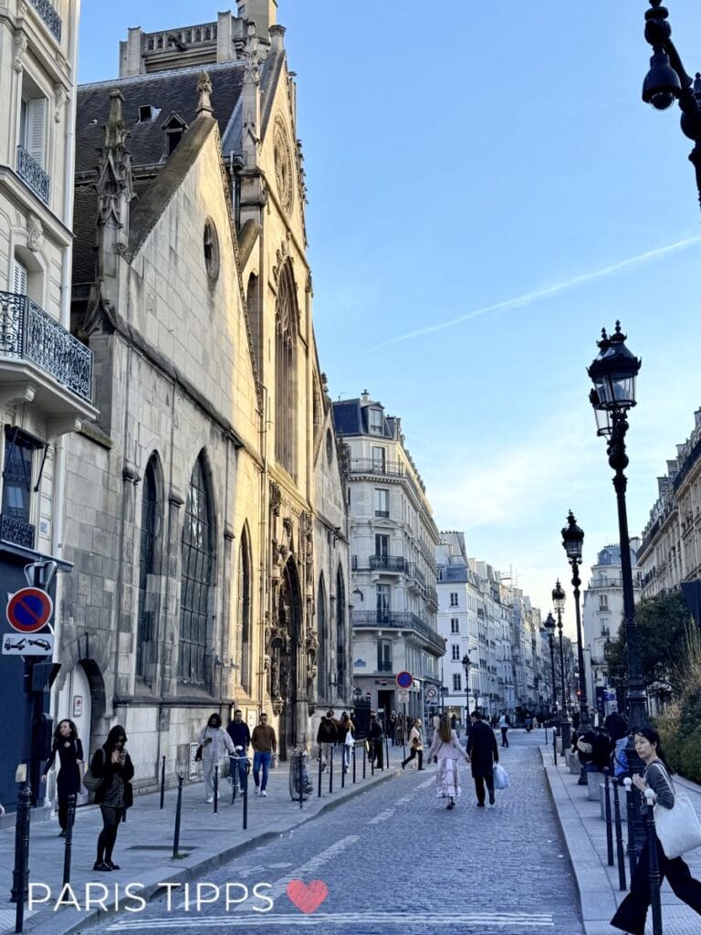 Ruhige Pariser Straße mit historischer Kirche, eleganten Altbauten und klassischen Laternen im warmen Abendlicht, während Menschen entspannt entlanggehen. Der Schriftzug „PARIS TIPPS ❤️“ passt zu einer zentralen und charmanten Umgebung für eine Paris mit Kindern Unterkunft.