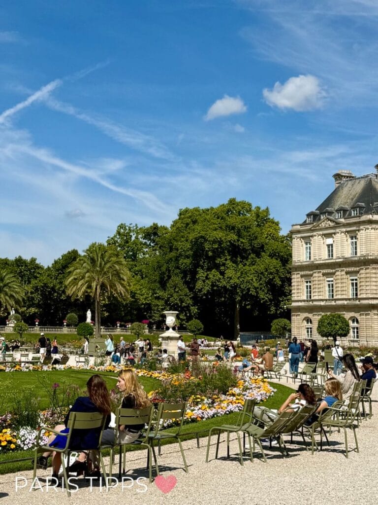 Menschen entspannen auf grünen Stühlen rund um bunte Blumenbeete in einem sonnigen Pariser Garten mit historischem Gebäude und hohen Bäumen im Hintergrund. Der Text "PARIS TIPPS ❤️" verweist auf Empfehlungen für Paris mit Kindern Unterkunft während Familien und Besucher die Anlage genießen.