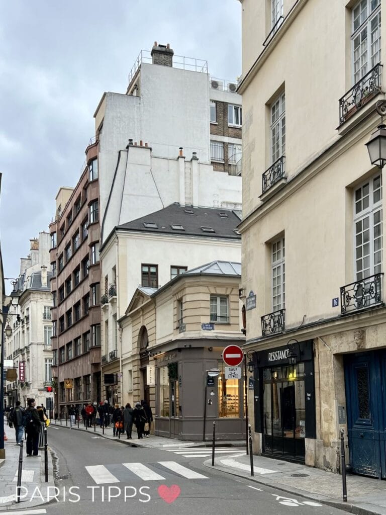 Schmale Pariser Straße mit klassischen Wohnhäusern, kleinen Boutiquen und Menschen, die entspannt entlanggehen, unter grauem Himmel. Der Schriftzug „PARIS TIPPS ❤️“ zeigt eine typische, ruhigere Wohngegend, die sich gut für eine Paris mit Kindern Unterkunft eignet.