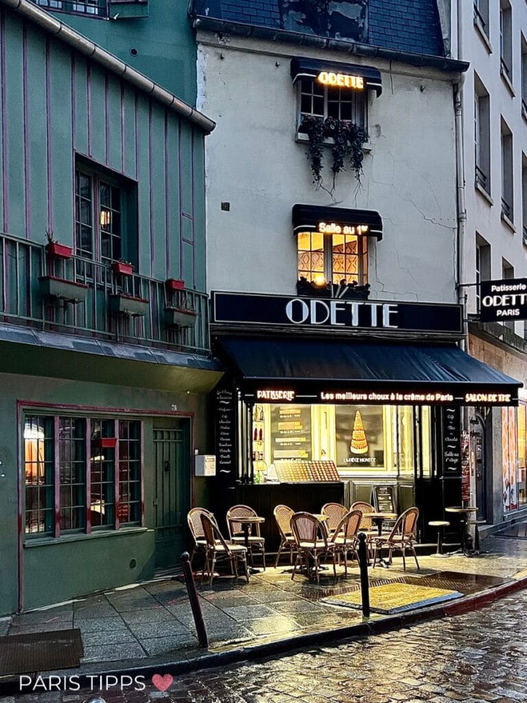 Odette Bäckerei im warmen Licht. Die Strasse ist Regennass, und Stühle und Tische stehen vor dem beleuchteten Schaufenster. Es ist dämmrig, fast dunkel.