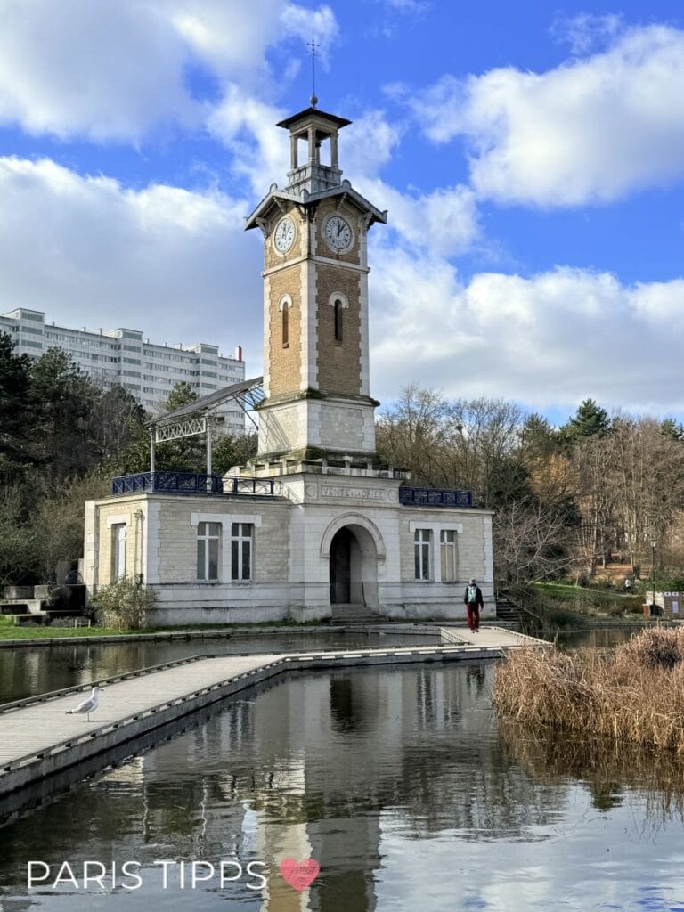 Kleiner Turm mit Uhr auf einem Gebäude am Wasser, umgeben von Natur und einem Holzsteg, auf dem Menschen spazieren gehen, unter blauem Himmel mit Wolken. Der Schriftzug „PARIS TIPPS ❤️“ passt zu einer ruhigeren Gegend abseits des Trubels für eine Paris mit Kindern Unterkunft.