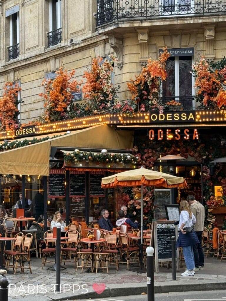 Alt text: Belebtes Pariser Café „ODESSA“ mit orangefarbenen Blumen, Lichterketten und Sitzplätzen im Freien vor einem klassischen Altbau in Montparnasse. Der Schriftzug „PARIS TIPPS ❤️“ passt zu Inspirationen für Paris mit Kindern Unterkunft in lebendiger, zentraler Lage mit Restaurants und Cafés.