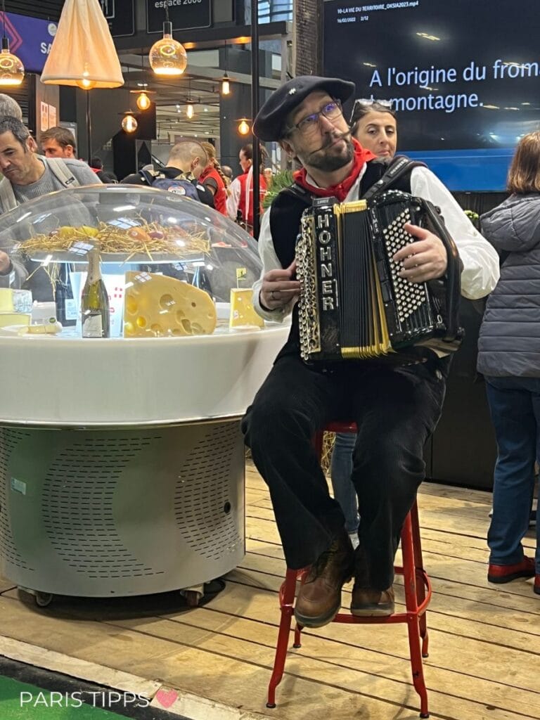Ein Mann mit Baskenmütze sitzt auf einem Hocker und spielt Akkordeon. Neben ihm eine Käseglocke, unter der sich die Produkte der Region befindent: Käse, Wein, Obst. Um ihn herum stehen Menschen, die den Salon besuchen.