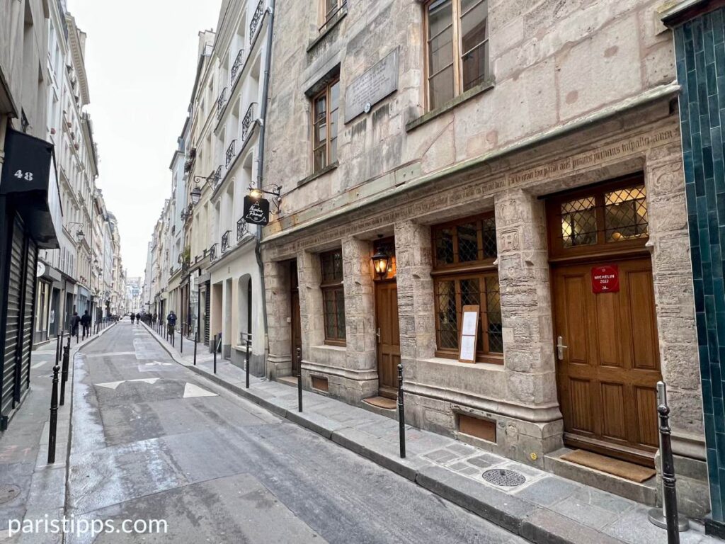 Eine schmale Pariser Straße mit historischen Gebäuden auf beiden Seiten. Auf der rechten Seite befindet sich ein altes Steinhaus mit geschnitzten Details, Holztüren und Fenstern, sowie einer roten Plakette mit dem Michelin-Stern. Links führen weitere moderne Gebäude und Laternen die ruhige, leicht nasse Straße entlang