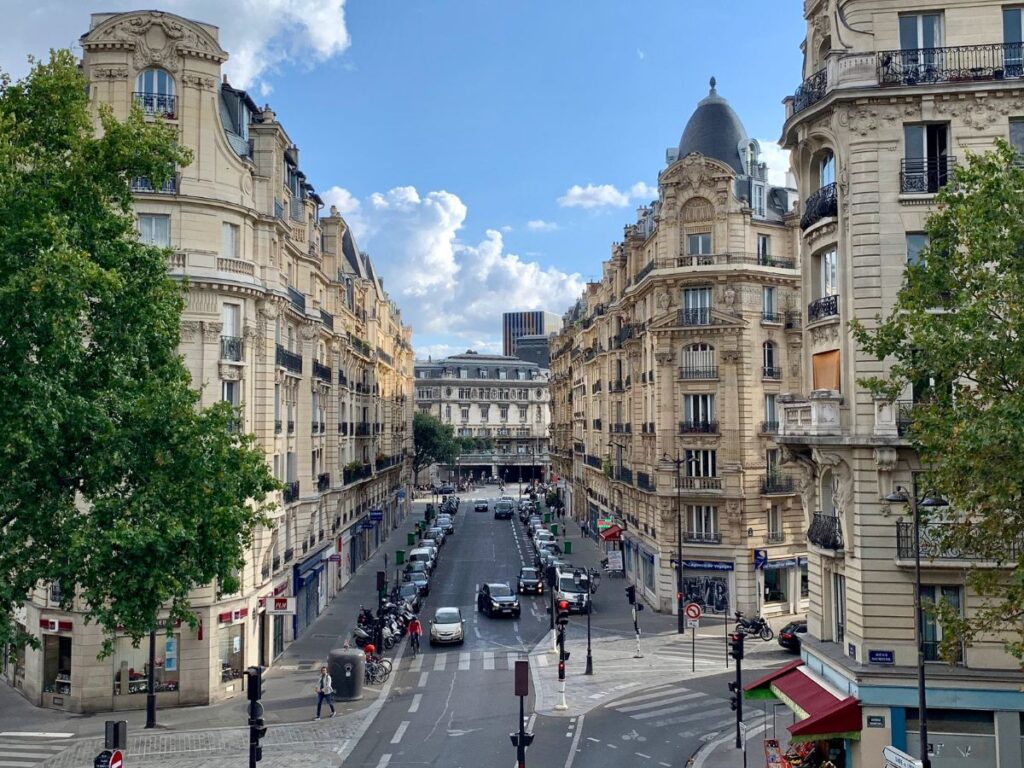 Elegante Pariser Wohngebäude mit aufwendig verzierten Fassaden säumen eine symmetrische Straße. Im Vordergrund sind grüne Bäume zu sehen, während der Himmel blau ist und von weißen Wolken durchzogen wird. Autos und Fußgänger beleben die Szene, die einen klassischen, gehobenen Pariser Stadtteil darstellt.