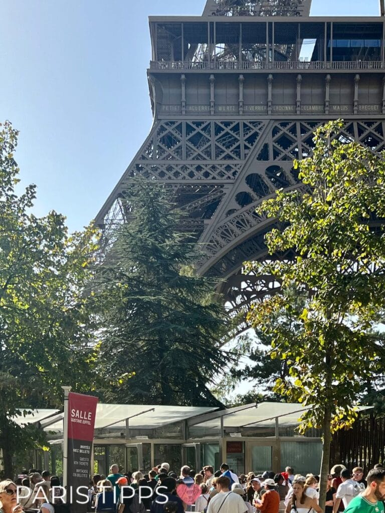 Menschenmenge am Eingang der 'Salle Gustave Eiffel' am Fuße des Eiffelturms, umgeben von Bäumen und bei strahlendem Sonnenschein. Im Hintergrund sind die beeindruckenden Metallstrukturen des Eiffelturms zu sehen
