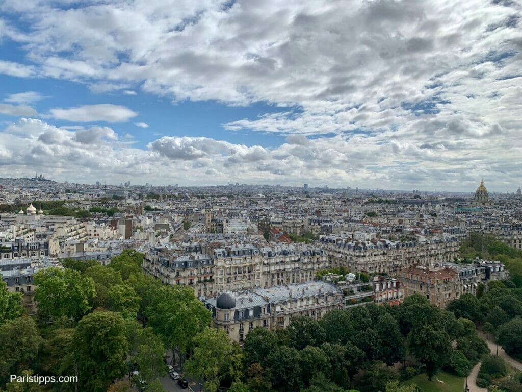 Aussicht von der Plattform der 2. Etage des Eiffelturms auf Paris, mit historischen Gebäuden, grünen Parks und einer weiten Sicht bis zum Horizont. Der Himmel ist teils bewölkt, teils blau, und Sehenswürdigkeiten wie die Basilika Sacré-Cœur links und der Invalidendom mit seiner goldenen Kuppel rechts sind erkennbar.