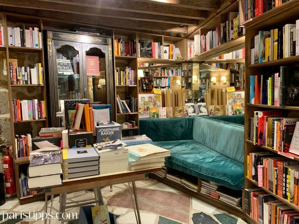 Ein gemütlicher Raum in der Buchhandlung 'Shakespeare and Company', mit Regalen voller Bücher, einem Tisch mit gestapelten Büchern und einer türkisfarbenen Samtcouch in der Ecke. Im Hintergrund sind ein verspiegeltes Regal und warme Wandlampen zu sehen, die dem Raum eine einladende Atmosphäre verleihen.