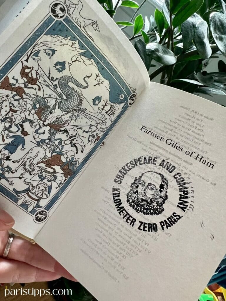 Eine aufgeschlagene Buchseite mit einer kunstvoll illustrierten Szene links, die Drachen und mittelalterliche Figuren zeigt, und rechts der Titel 'Farmer Giles of Ham' sowie ein runder Stempel mit der Aufschrift 'Shakespeare and Company Kilometer Zero Paris' und dem Porträt von Shakespeare. Im Hintergrund sind grüne Pflanzen sichtbar.