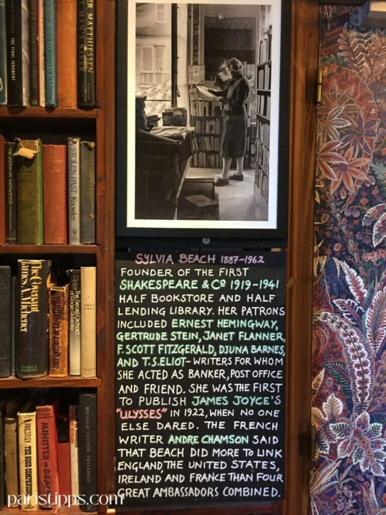 Eine Wand in der Buchhandlung 'Shakespeare and Company', dekoriert mit einem Regal voller Bücher, einer Schwarz-Weiß-Fotografie von Sylvia Beach in ihrer Buchhandlung und einer Texttafel. Der Text beschreibt Sylvia Beach (1887–1962) als Gründerin von Shakespeare & Co. und erwähnt berühmte Autoren wie Hemingway, Fitzgerald und Joyce, mit denen sie zusammenarbeitete