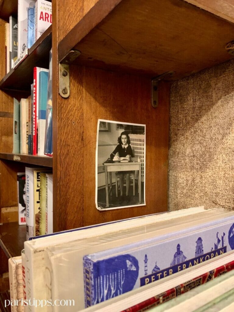 Ein Regal in der Buchhandlung 'Shakespeare and Company', mit Büchern und einem Schwarz-Weiß-Foto von Anne Frank, das an die hölzerne Regalwand geklebt wurde. Die Bücher im Vordergrund umfassen verschiedene Titel, darunter 'Peter Frankopan', und die Szene strahlt eine historische und nachdenkliche Atmosphäre aus."