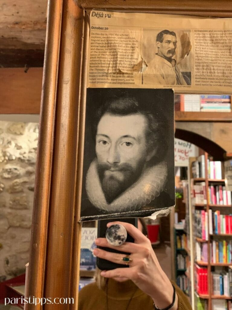 Ein vergoldeter Spiegel in der Buchhandlung 'Shakespeare and Company', dekoriert mit einer Schwarz-Weiß-Abbildung eines historischen Porträts und einem Zeitungsausschnitt mit der Überschrift 'Déjà vu'. Im Spiegel ist die Hand einer Person mit einem Smartphone sichtbar, das ein Bild der Szene aufnimmt. Im Hintergrund sind Bücherregale zu erkennen.