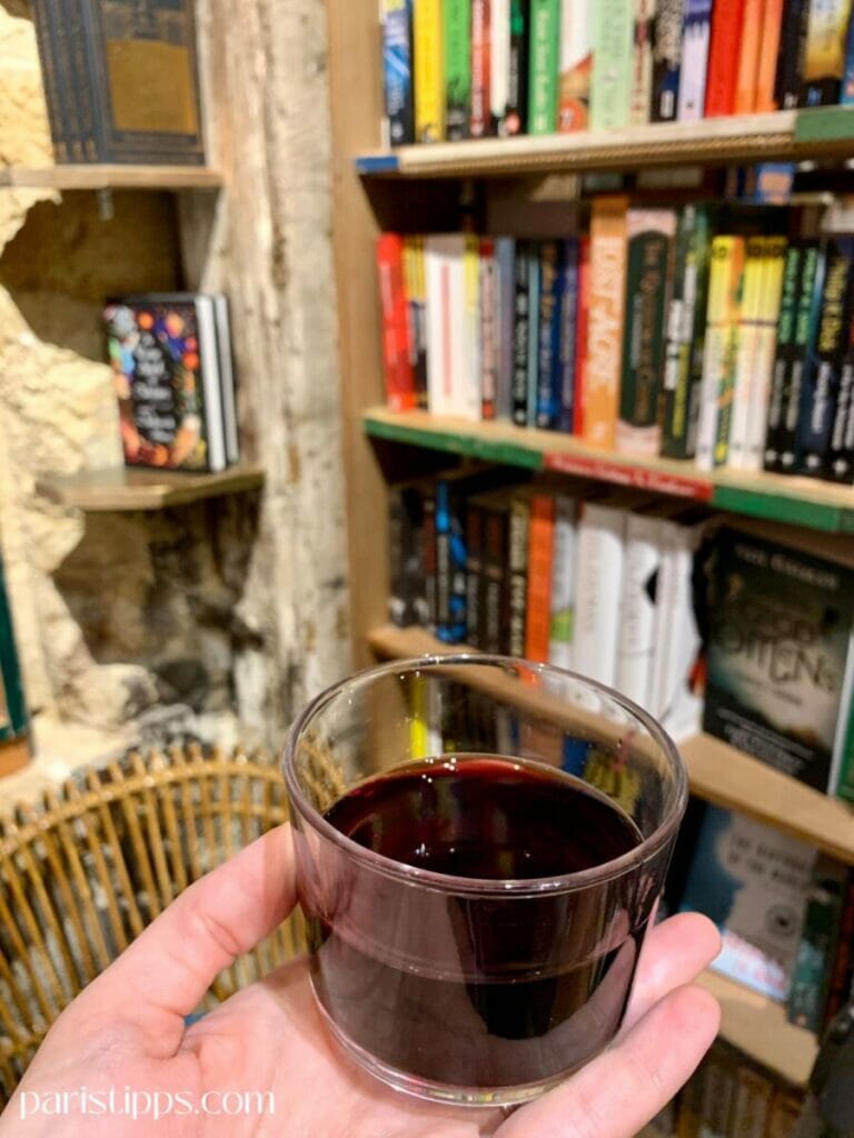 Eine Hand hält ein Glas mit Rotwein vor einem Bücherregal in der Buchhandlung 'Shakespeare and Company'. Im Hintergrund sind bunt gemischte Bücher sowie ein Rattanstuhl und eine Wand aus hellem Stein zu sehen, die eine warme, einladende Atmosphäre schaffen