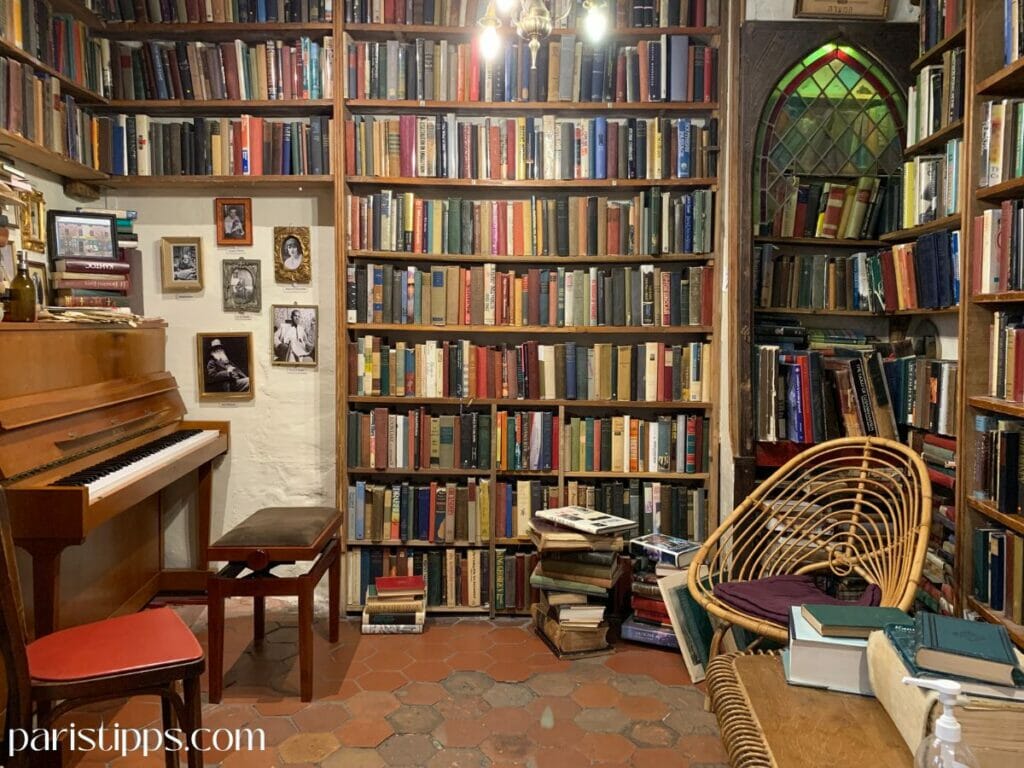 Ein gemütlicher Raum in der Buchhandlung 'Shakespeare and Company', gefüllt mit deckenhohen Bücherregalen, einem Klavier mit Hocker und gerahmten Schwarz-Weiß-Fotografien an der Wand. Im Vordergrund stehen ein Rattansessel und ein Tisch mit Büchern, während ein buntes Buntglasfenster im Hintergrund eine warme Atmosphäre schafft.
