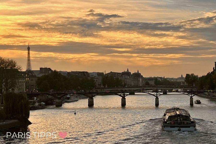 Ein Ausflugsboot fährt bei Sonnenuntergang über die Seine mit Blick auf den beleuchteten Eiffelturm und eine Brücke voller Menschen. Der Himmel ist wolkig mit warmem Licht am Horizont und spiegelt sich im Wasser. Unten links steht "PARIS TIPPS " und vermittelt die Stimmung einer abendlichen Bootsfahrt in Paris.