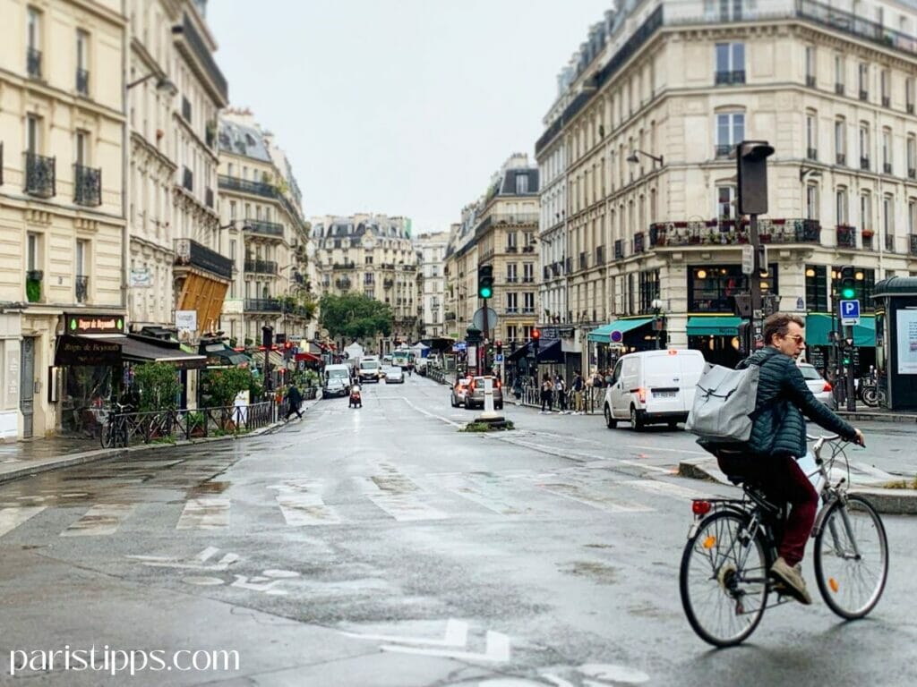 Radfahrer fährt durch eine nasse Straße in Paris bei Regen. Die Szene zeigt typische Verkehrsbedingungen und verdeutlicht, dass Radfahren in Paris auch bei schlechtem Wetter stattfindet. Erfahrung im Stadtverkehr ist hier besonders wichtig.