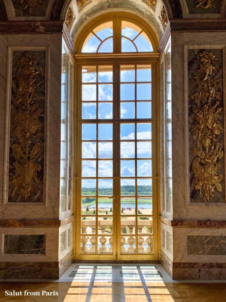Prunkvolles Fenster mit goldenen Verzierungen und Blick auf Gärten von Versailles und blauen Himmel mit dem Text "Salut from Paris". Diese Szene spiegelt die Eleganz der Paris Sehenswürdigkeiten wider.