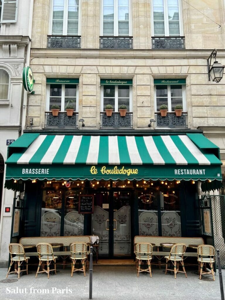 "Fassade eines Pariser Cafés namens 'Le Bouledogue' mit einer grün-weiß gestreiften Markise. Darunter stehen Tische und grün gestreifte Stühle vor der Glasfront des Cafés, das von Pflanzen und charmanten Fenstern mit Topfpflanzen eingerahmt wird.