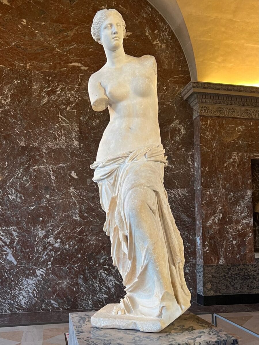Weiße Marmorskulptur der Venus von Milo im Louvre, dargestellt ohne Arme vor einer dunklen Marmornische. Die Statue steht auf einem Sockel und ist eines der bekanntesten Werke nach dem eintritt louvre.