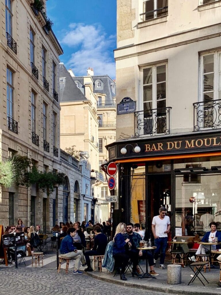 Straße in Paris mit Straßencafé und Leuten, die draussen an Tischen sitzen. 