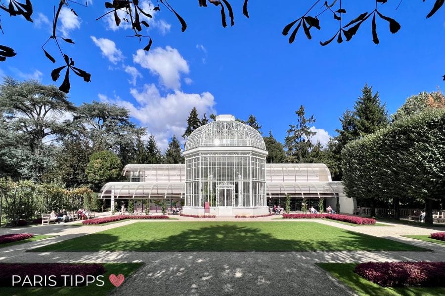 Orangerie vor einer grünen Wiese und blauem Himmel. Links und rechts sind Bäume und Bänke, auf denen Menschen in der Sonne sitzen.