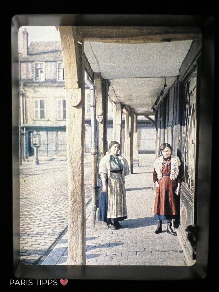 Zwei Frauen in traditioneller Kleidung stehen unter einer überdachten Passage in einer historischen Straßenszene.