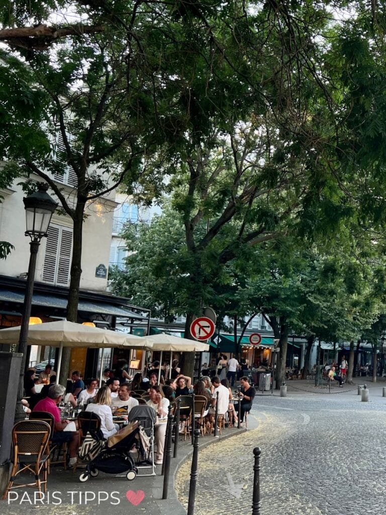 Belebtes Straßencafé auf einem kopfsteingepflasterten Platz in Montmartre, umgeben von grünen Bäumen und entspannter Pariser Atmosphäre.