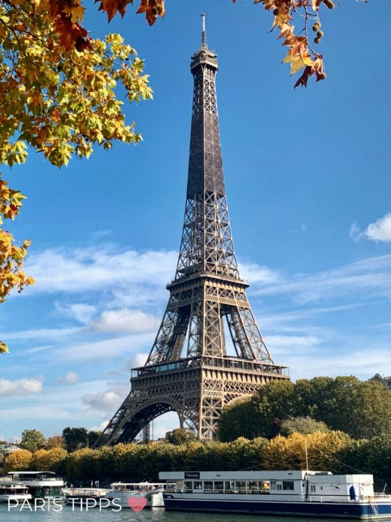 Eiffelturm umgeben von goldenem Herbstlaub und blauem Himmel. Im Vordergrund die Seine mit Ausflugsschiffen.